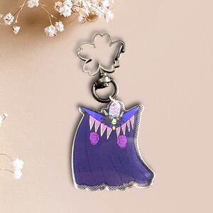 May include: A purple and pink anime character keychain with a white and black striped hat. The character is wearing a purple cape with pink accents and has a black and white face.
