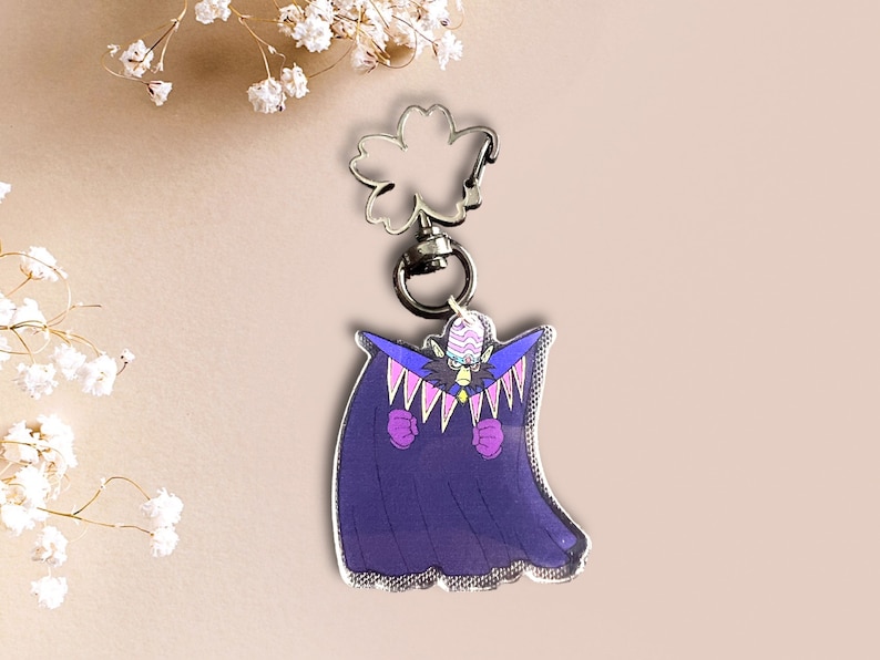 May include: A purple and pink anime character keychain with a white and black striped hat. The character is wearing a purple cape with pink accents and has a black and white face.