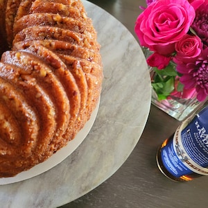 May include: A golden brown bundt cake with pecan pieces on top, sitting on a white plate on a marble surface. A bottle of Basil Hayden's Kentucky Straight Bourbon Whiskey with a blue label and a gold cap is in the background.