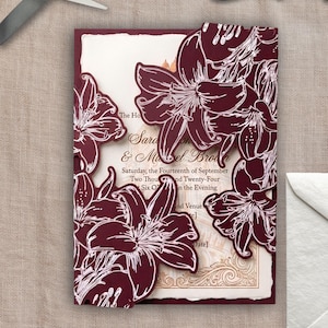 May include: A wedding invitation card featuring deep burgundy die-cut lily floral overlays on a cream-colored background with gold-toned text reading Sarah and Michael Brown, Saturday, the Fourteenth of September, Two Thousand and Twenty-Four.