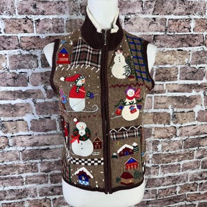 Op de afbeelding: Bruin vest met ritssluiting met een winterthema. Het vest heeft geborduurde sneeuwpoppen, huizen en ruitpatronen. Het heeft een crèmekleurige kraag en een volledige ritssluiting. Het vest wordt getoond op een mannequin.