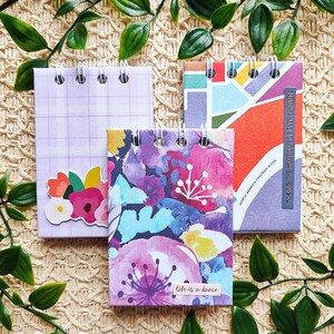 Könnte beinhalten: Drei kleine spiralgebundene Notizbücher mit verschiedenen Designs. Das erste Notizbuch hat einen lilafarbenen Einband mit einem Gittermuster und einem Blumenaufkleber. Das zweite Notizbuch hat ein Aquarell-Blumendesign mit dem Text "Life is a dance". Das dritte Notizbuch hat ein geometrisches Design mit dem Text "Having the most fun in the whole wide world".