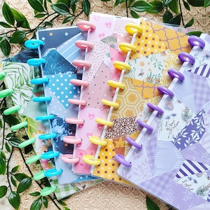 Pode incluir: Uma coleção de cadernos coloridos com encadernação de discos de plástico. As capas apresentam designs de patchwork em várias cores, incluindo rosa, amarelo e roxo. Os cadernos estão dispostos em uma exibição em camadas.