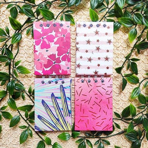 Op de afbeelding: Vier spiraalgebonden notitieboekjes met kleurrijke ontwerpen. De notitieboekjes hebben bloemen-, sterren-, palmblad- en abstracte patronen in roze, blauw en geel. De notitieboekjes zijn gerangschikt op een geweven oppervlak met groen gebladerte.