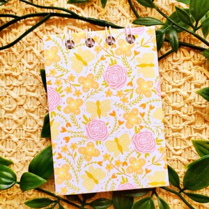 May include: A small white notebook with a floral pattern featuring pink roses, yellow butterflies, and orange flowers. The notebook has a spiral binding and is lying on a woven straw surface.