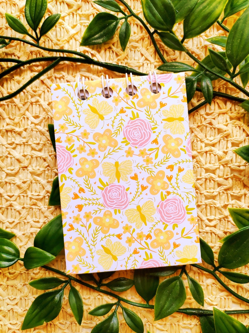 May include: A small white notebook with a floral pattern featuring pink roses, yellow butterflies, and orange flowers. The notebook has a spiral binding and is lying on a woven straw surface.