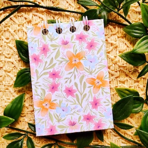 May include: A small spiral bound notebook with a floral pattern. The cover features a white background with pink, orange, and blue flowers and green leaves.