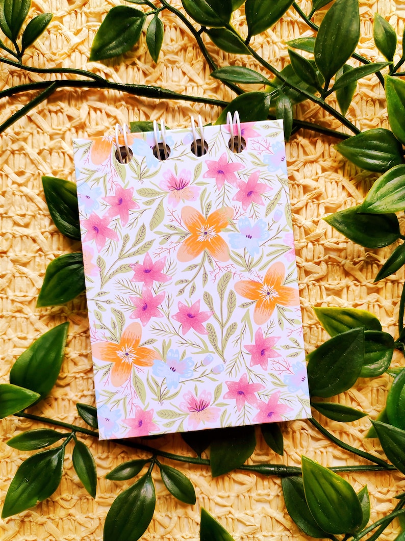 May include: A small spiral bound notebook with a floral pattern. The cover features a white background with pink, orange, and blue flowers and green leaves.