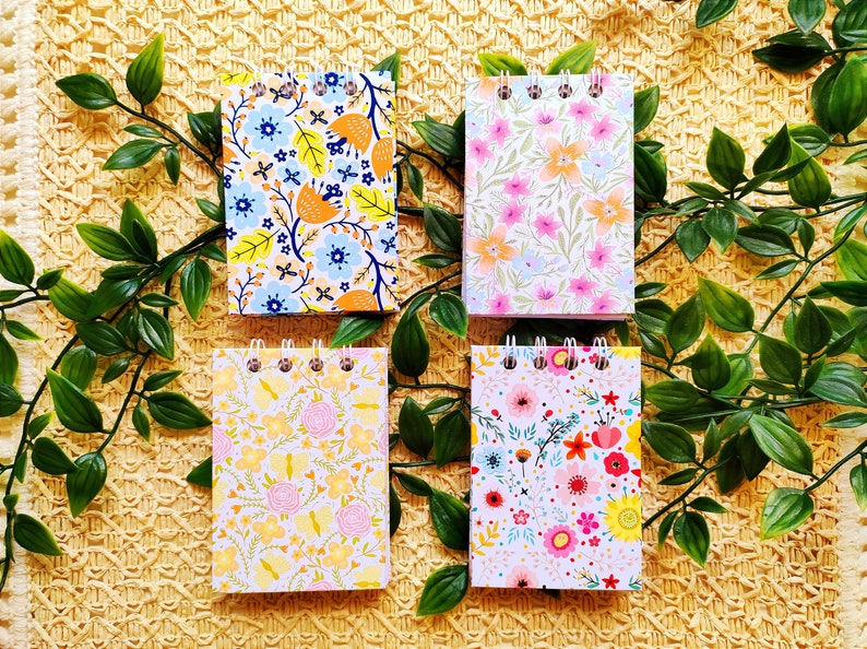 May include: Four small spiral bound notebooks with floral patterns. The notebooks have white covers with different colored floral designs. The notebooks are on a yellow woven surface.