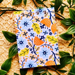 May include: A small spiral bound notebook with a white background and a colorful floral pattern. The pattern includes orange, yellow, blue, and green flowers and leaves.