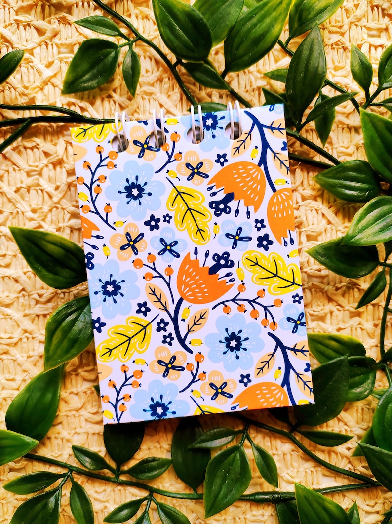 May include: A small spiral bound notebook with a white background and a colorful floral pattern. The pattern includes orange, yellow, blue, and green flowers and leaves.