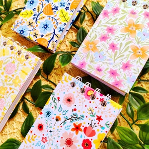 May include: Four small spiral bound notebooks with floral patterns. The notebooks have white covers with different colored flowers and leaves. The notebooks are on a yellow surface with green leaves.
