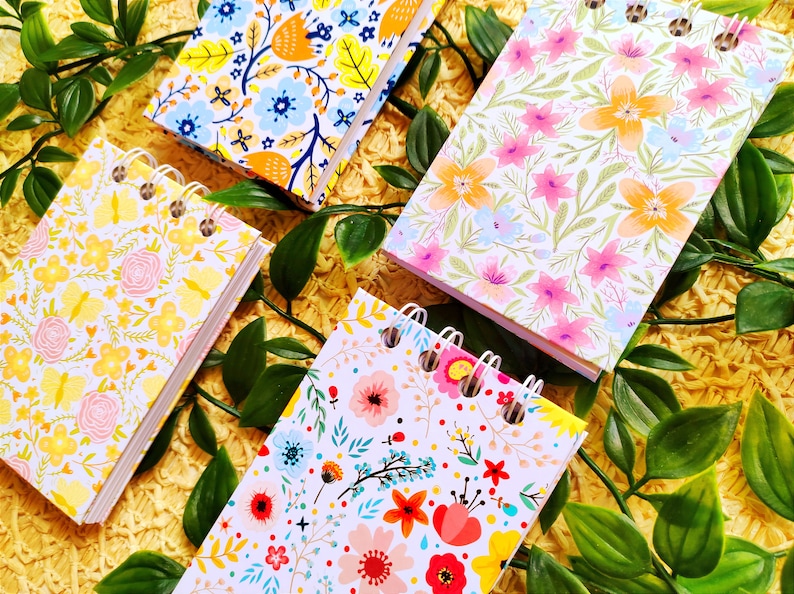 May include: Four small spiral bound notebooks with floral patterns. The notebooks have white covers with different colored flowers and leaves. The notebooks are on a yellow surface with green leaves.