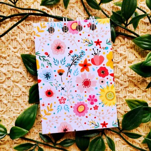 May include: A small spiral bound notebook with a white background and a colorful floral pattern. The notebook is surrounded by green leaves.