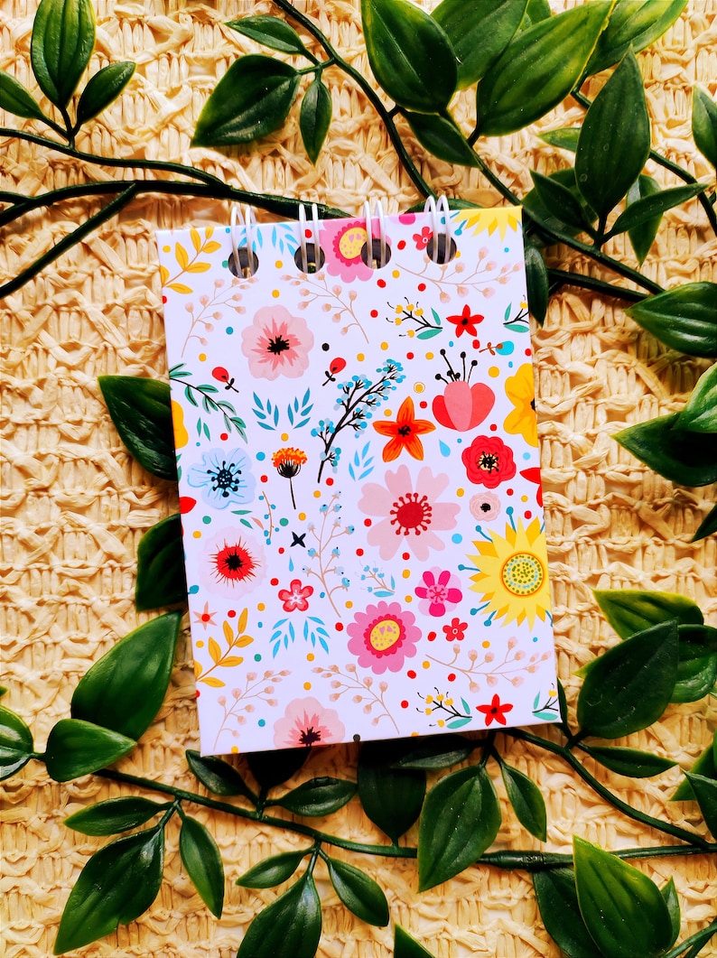 May include: A small spiral bound notebook with a white background and a colorful floral pattern. The notebook is surrounded by green leaves.