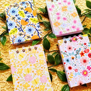 May include: Four small spiral bound notebooks with floral patterns. The notebooks have white backgrounds with different colored flowers and leaves. The notebooks are on a yellow woven surface.