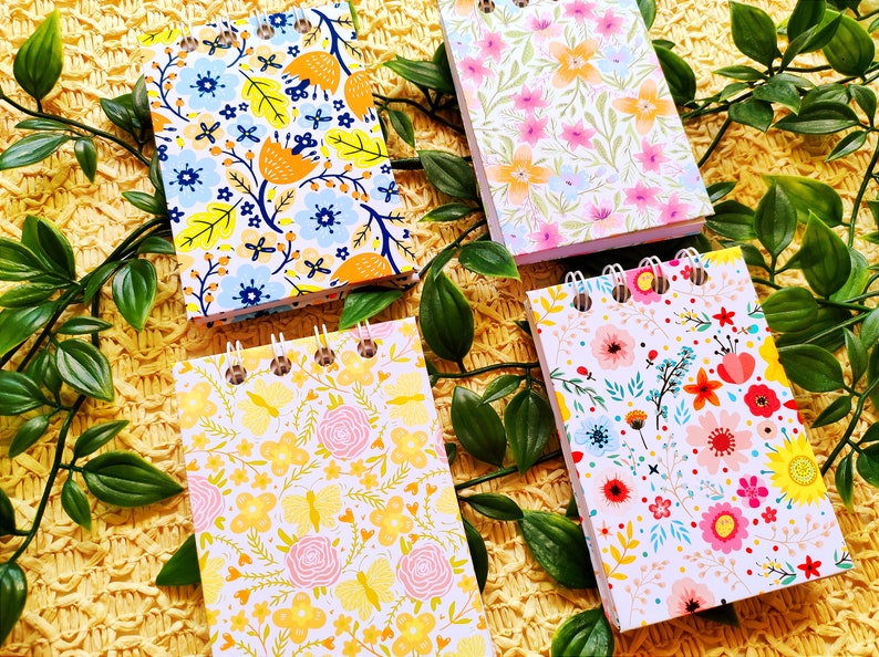 May include: Four small spiral bound notebooks with floral patterns. The notebooks have white backgrounds with different colored flowers and leaves. The notebooks are on a yellow woven surface.