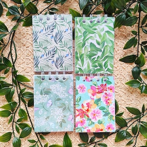 Op de afbeelding: Vier spiraalgebonden notitieboekjes met bloemen- en botanische ontwerpen. De notitieboekjes hebben patronen van groene bladeren, blauwe vogels en kleurrijke bloemen. De notitieboekjes zijn gerangschikt op een geweven achtergrond met groen gebladerte.