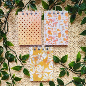 May include: Three spiral-bound notepads with patterned covers. One has orange polka dots, another has a repeating abstract design in orange and pink, and the third features white daisies on a yellow background. Green leafy vines surround the notepads.