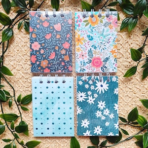 Puede incluir: Cuatro cuadernos con espiral con patrones florales y de puntos. Los cuadernos presentan una variedad de diseños, incluyendo un patrón floral azul oscuro con flores rosas y naranjas, un arreglo floral colorido, un patrón de puntos azul claro y un patrón floral turquesa con margaritas blancas.