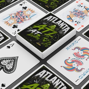 May include: A deck of playing cards featuring a central card with an Atlanta-themed graphic, including an alien wearing a hat, the text ATLANTA, and green brush strokes. Surrounding cards display traditional king, joker, and ace of spades designs.