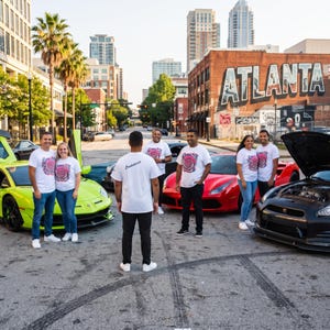 May include: A group of people wearing white t-shirts with a circular graphic design stand on a city street among several sports cars, including a lime green Lamborghini, a red Ferrari, and a black Nissan GT-R with its hood open.