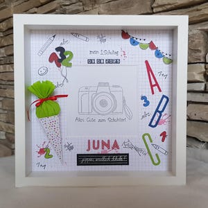 Könnte beinhalten: Ein weiß gerahmtes Kunstwerk mit Schulthema. Es zeigt eine Kamera-Illustration, den Namen "JUNA" und das Datum "09 08 2025". Dekorative Elemente sind eine Papiertüte, Luftballons sowie bunte Buchstaben und Zahlen. Der Text "Alles Gute zum Schulstart" ist ebenfalls vorhanden.