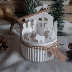 Könnte beinhalten: Weiße Holz-Krippe mit einem Stern oben, Figuren von Maria, Josef, Jesuskind und einem Schaf. Die Szene befindet sich in einer runden Basis mit braunem Seilbesatz und dem Text "Gott ist Liebe" auf einem Etikett.