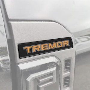 Può includere: Primo piano di un pannello di veicolo argentato con un emblema "TREMOR" nero e arancione. L'emblema è rettangolare con la parola "TREMOR" in lettere arancioni. Il design del veicolo è visibile sullo sfondo.