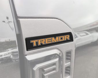 May include: Close-up of a silver vehicle panel with a black and orange "TREMOR" emblem. The emblem is rectangular with the word "TREMOR" in orange lettering. The vehicle's design is visible in the background.