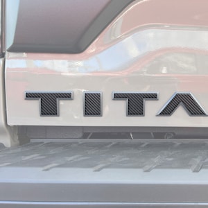 May include: A close-up of a silver truck tailgate with a black carbon fiber "TITAN" emblem.