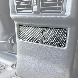 Puede incluir: Un panel de fibra de carbono con dos puertos USB, instalado en el interior de un vehículo. El panel tiene un patrón tejido en negro y gris, y está situado debajo de las rejillas de ventilación.