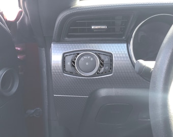 May include: A close-up of a car's interior, showing a black and silver dashboard with a carbon fiber trim. The dashboard features a round control knob with multiple buttons and a silver ring around it. The control knob is labeled "RD" and "LS".