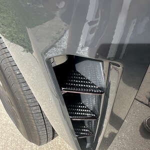 Puede incluir: Primer plano del parachoques delantero de un coche con una inserci&oacute;n de ventilaci&oacute;n de fibra de carbono negra. El parachoques es de color gris oscuro y el neum&aacute;tico es visible en el lado izquierdo. La inserci&oacute;n de ventilaci&oacute;n tiene un dise&ntilde;o triangular.