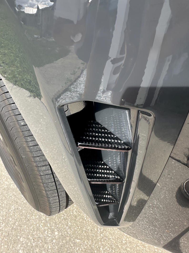 Puede incluir: Primer plano del parachoques delantero de un coche con una inserci&oacute;n de ventilaci&oacute;n de fibra de carbono negra. El parachoques es de color gris oscuro y el neum&aacute;tico es visible en el lado izquierdo. La inserci&oacute;n de ventilaci&oacute;n tiene un dise&ntilde;o triangular.