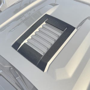 May include: A close-up of a car hood with a rectangular vent. The vent has a carbon fiber trim and a series of horizontal slats. The hood is a light gray color, and the vent is a darker gray.