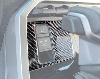 May include: Close-up of a black carbon fiber trim panel for a car interior. The panel features two buttons, one labeled "HOLD" and the other labeled "PULL ON (P)".