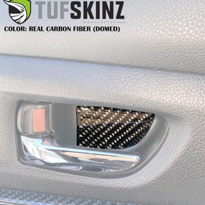 May include: A black and gray real carbon fiber (domed) insert for a car door handle. The insert is a geometric pattern and is set against a gray interior. The text "Tufskinz Manufacturing" and "Color: Real Carbon Fiber (Domed)" are visible.