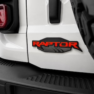 May include: Close-up of a white vehicle's rear with a black bumper and a red "RAPTOR" emblem. The emblem is outlined in black and features a mountain graphic. A portion of a black tire is visible on the right.