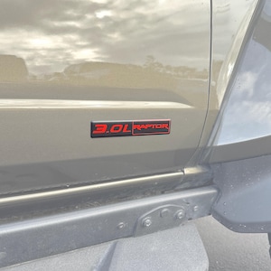May include: A close-up of a vehicle's side panel featuring a black and red emblem. The emblem reads "3.0L RAPTOR" in a bold, modern font. The vehicle's body is a muted olive green color.