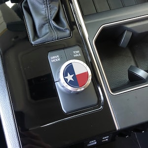 May include: A close-up of a car's interior showing a black and silver drive mode knob with a Texas flag design. The knob is labelled "Drive Mode" and "Tow/Haul".