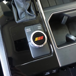 May include: Close-up of a black car's centre console with a silver knob labelled "Drive Mode" and "Tow/Haul". The knob has a red, yellow and black logo.
