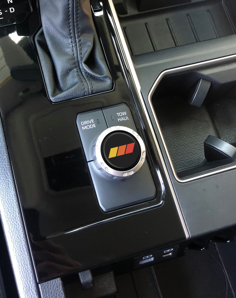 May include: Close-up of a black car's centre console with a silver knob labelled "Drive Mode" and "Tow/Haul". The knob has a red, yellow and black logo.