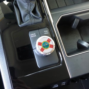 May include: Close-up of a car's centre console with a black leather gear shifter, a silver drive mode knob with the Florida state seal, and a black button labelled "Tow/Haul".