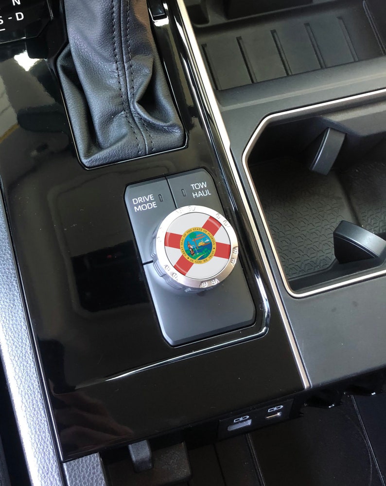 May include: Close-up of a car's centre console with a black leather gear shifter, a silver drive mode knob with the Florida state seal, and a black button labelled "Tow/Haul".