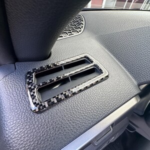 Puede incluir: Primer plano de una rejilla de ventilaci&oacute;n del salpicadero de un coche con un patr&oacute;n de fibra de carbono. La rejilla tiene forma rectangular con dos ranuras y un marco cromado. El salpicadero es negro y texturizado.