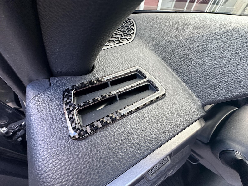 Puede incluir: Primer plano de una rejilla de ventilaci&oacute;n del salpicadero de un coche con un patr&oacute;n de fibra de carbono. La rejilla tiene forma rectangular con dos ranuras y un marco cromado. El salpicadero es negro y texturizado.