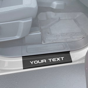 以下が含まれることがあります： カーボンファイバーのパターンと白い文字で「YOUR TEXT」と書かれた黒い車のドアシルです。