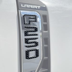 May include: Close-up of a silver vehicle panel with the word "LARIAT" at the top. Below, the letters "F-250" are prominently displayed in a textured, dark pattern. The panel has a reflective, chrome-like finish.