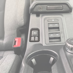 May include: A black carbon fiber trim kit for a car interior. The kit includes a switch panel with four buttons and a cup holder.
